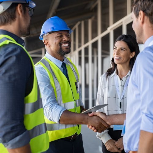 Smiling engineer shaking hands at construction site with happy architect. Handshake between cheerful african construction manager with businessman at bulding site. Team of workers with architects and contractor conclude an agreement with safety uniform.